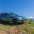 A green Subaru Outback SUV parked on a grassy dirt hillside with a mountain and blue sky in the background.
