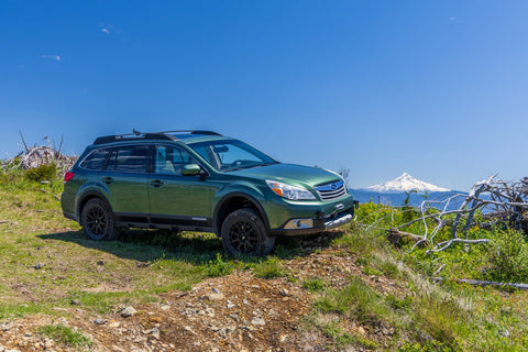 A green Subaru Outback SUV parked on a grassy dirt hillside with a mountain and blue sky in the background.