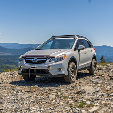 A white Subaru off-road Crosstrek parked on a rocky mountain trail with a scenic valley and blue sky in the background.